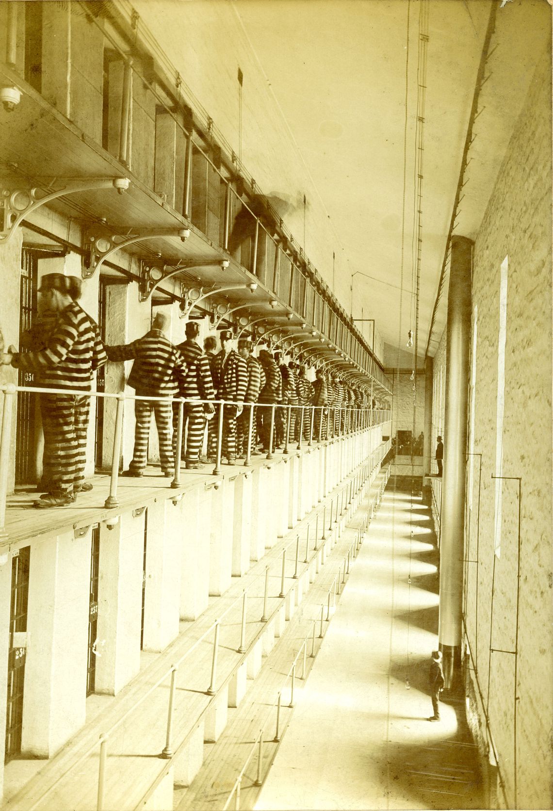 A sepia-toned photo shows a multitiered prison interior. On one of the upper floors, a number of people wearing black-and-white striped uniforms are standing in front of the cells. 