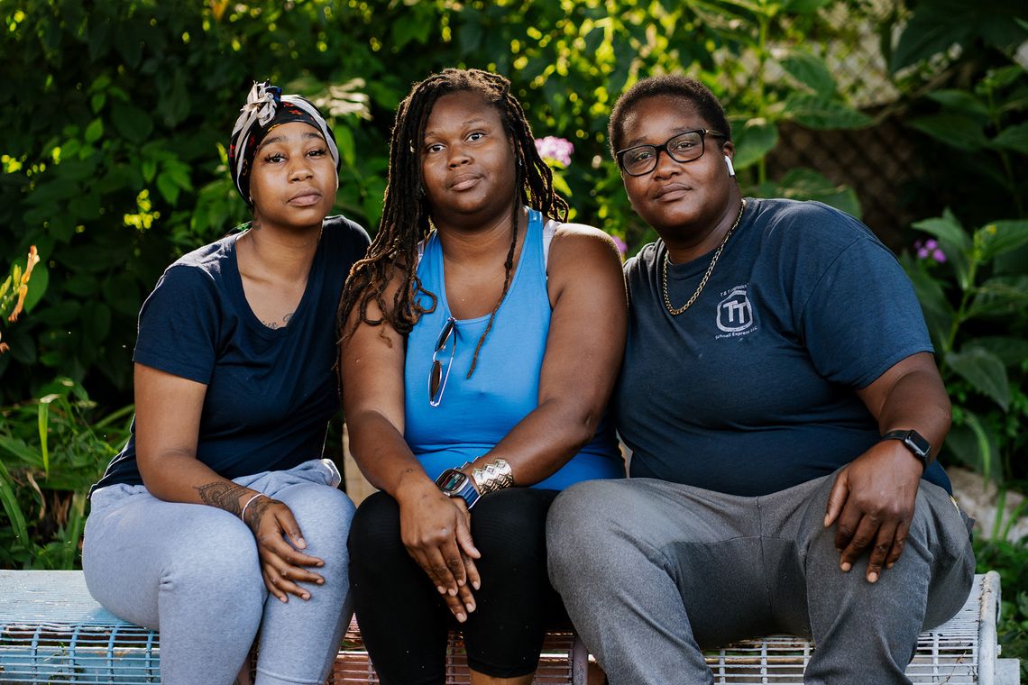 Three Black women sit on a white metal bench in a garden as they pose for a portrait.