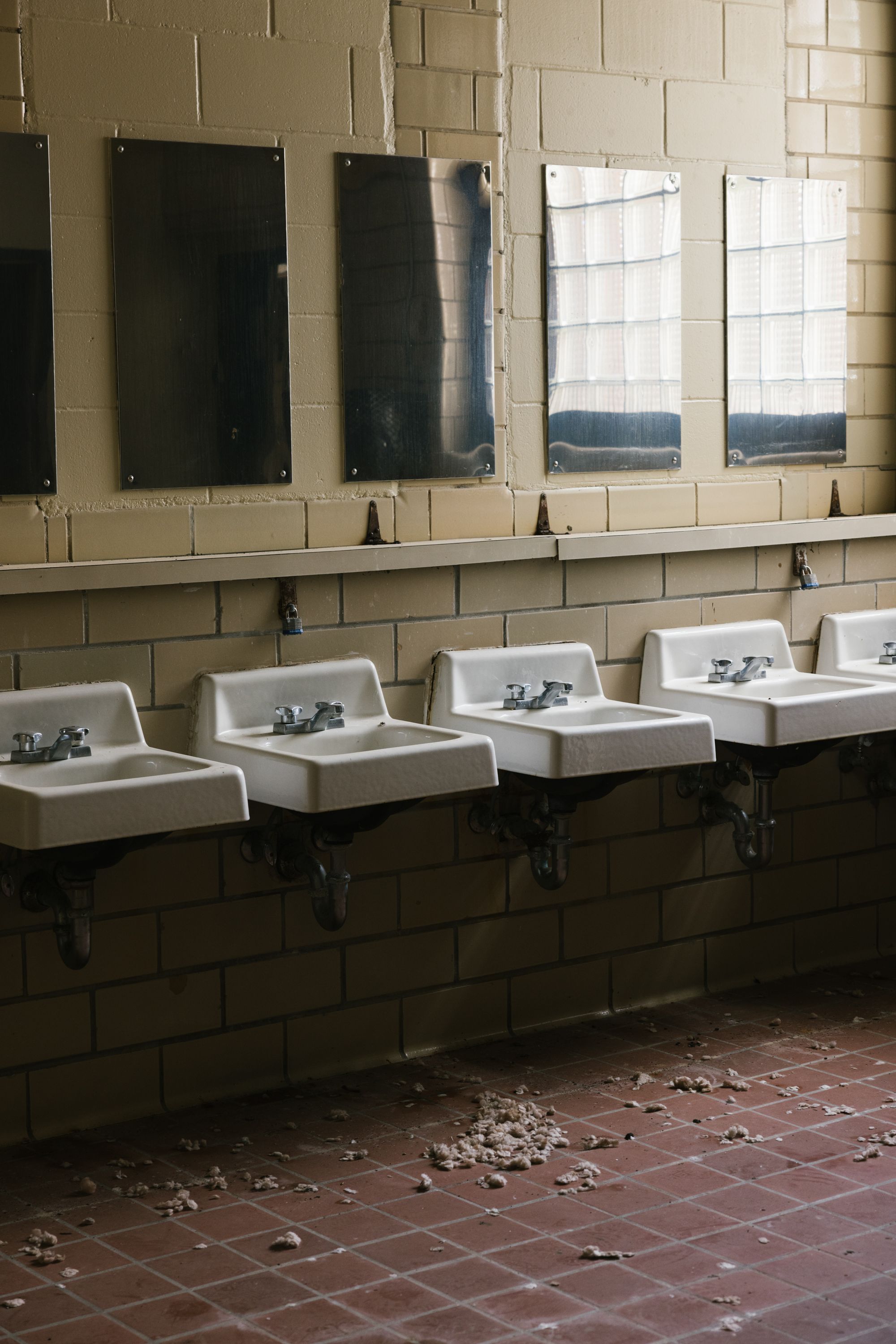 Five white sinks are visible in a bathroom that has metal mirrors above them. There is debris on the floor.