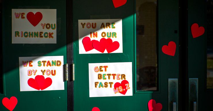 Signs and red paper hearts are taped to a green school door. The signs read as messages of support, such as: "We love you Richneck," "You are in our hearts," "We stand by you," and "Get better fast Ms. Z." 