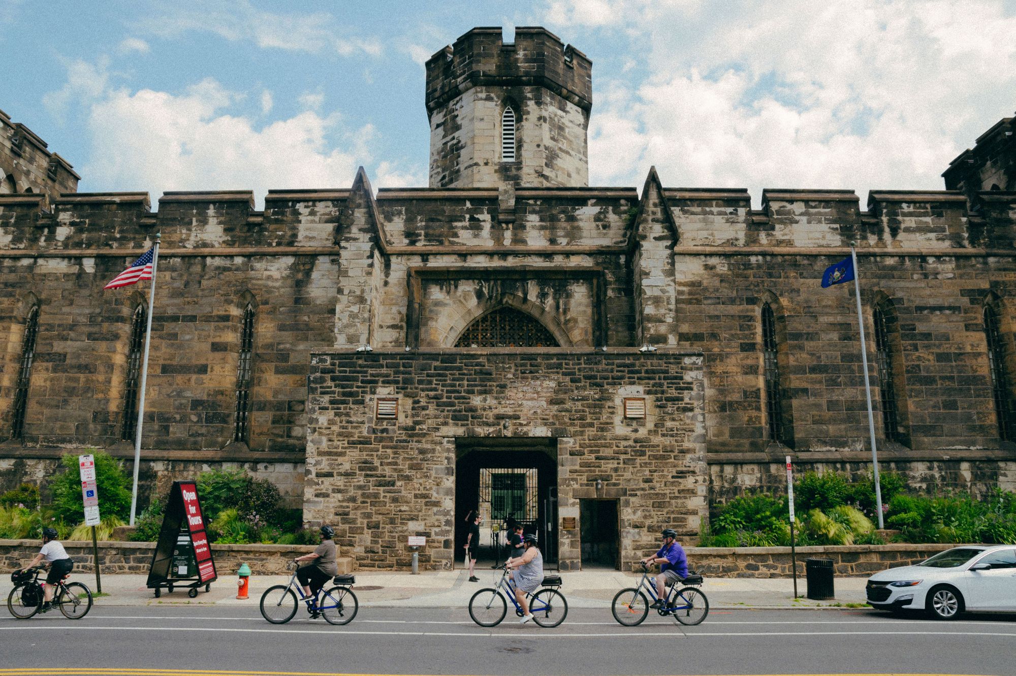 The exterior of Eastern State Penitentiary is visible, and looks like a stone castle. Four people ride bicycles in front of the doorway. 