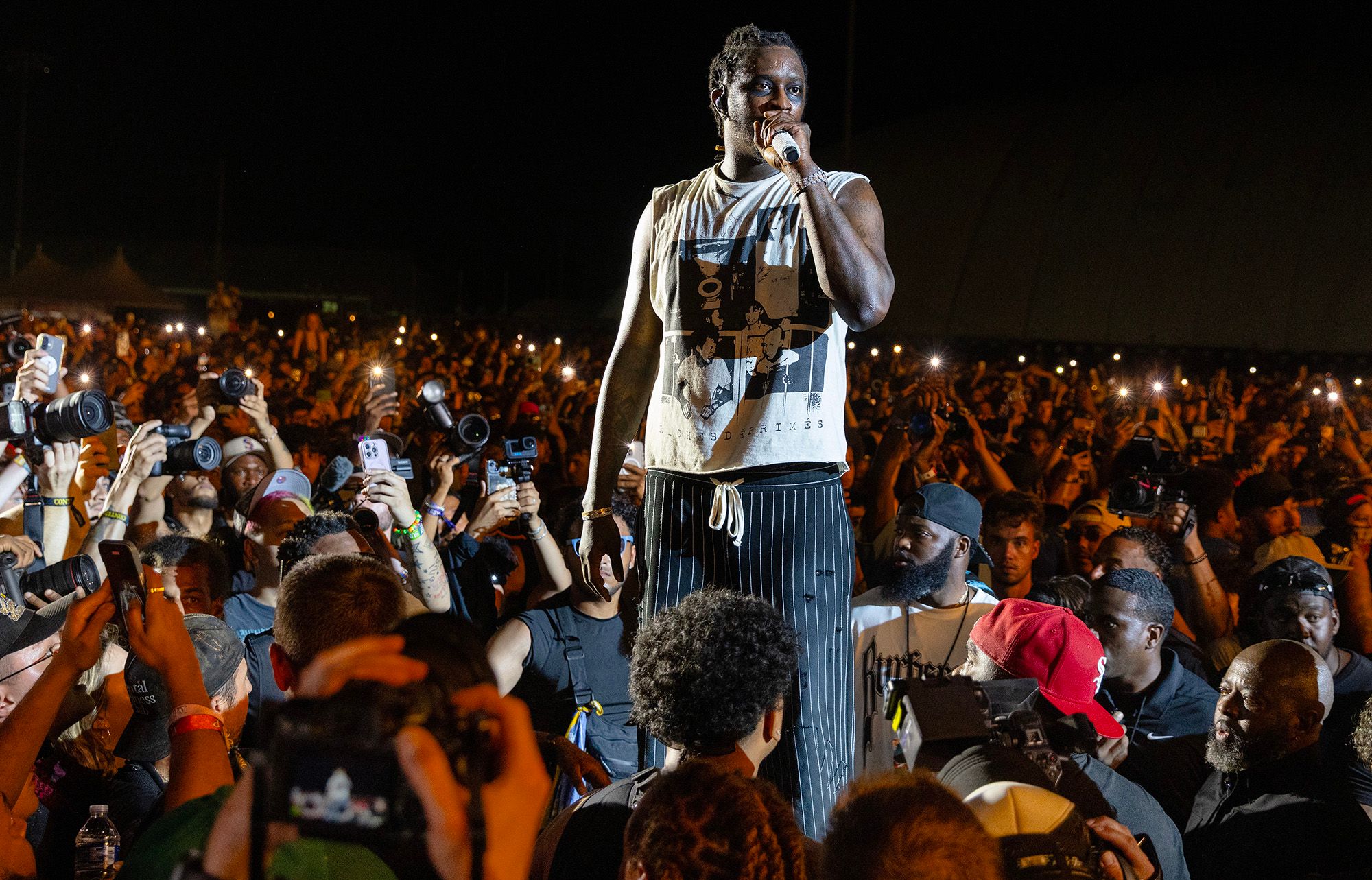 Young Thug, a Black rapper wearing a white shirt and striped pants, holds a mic as he performs in a crowd during a concert. 