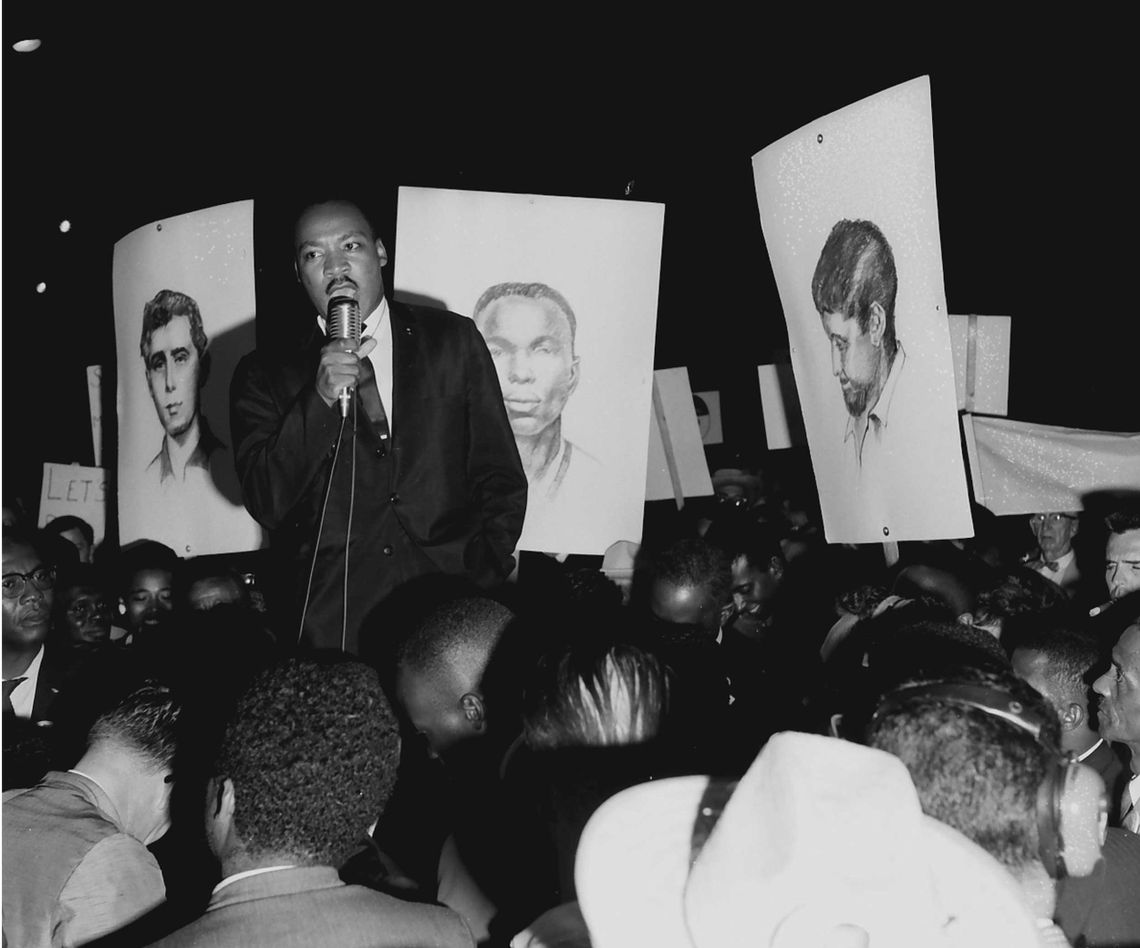 A black-and-white photo shows a Black man in a suit, Martin Luther King Jr., standing above a crowd of people while holding a microphone. Behind him are poster-sized sketches of two White men and one Black man. 