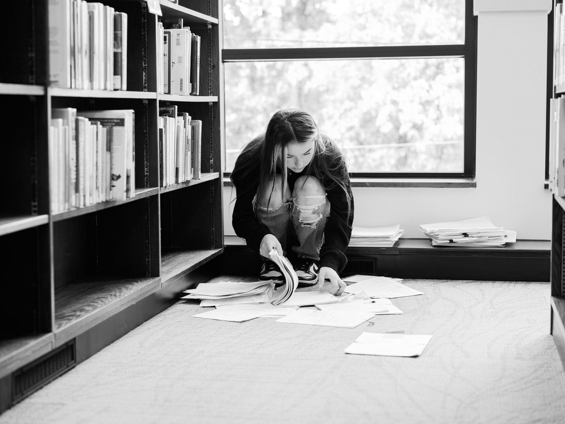 A black-and-white photo shows a White woman crouched down looking at paper records on the floor of a library. 