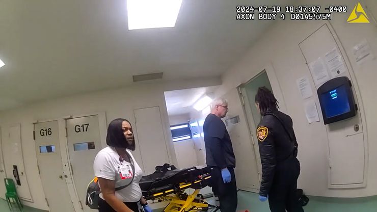 Three people stand outside a door in a jail. Behind a man with short white hair is an empty medical stretcher.  