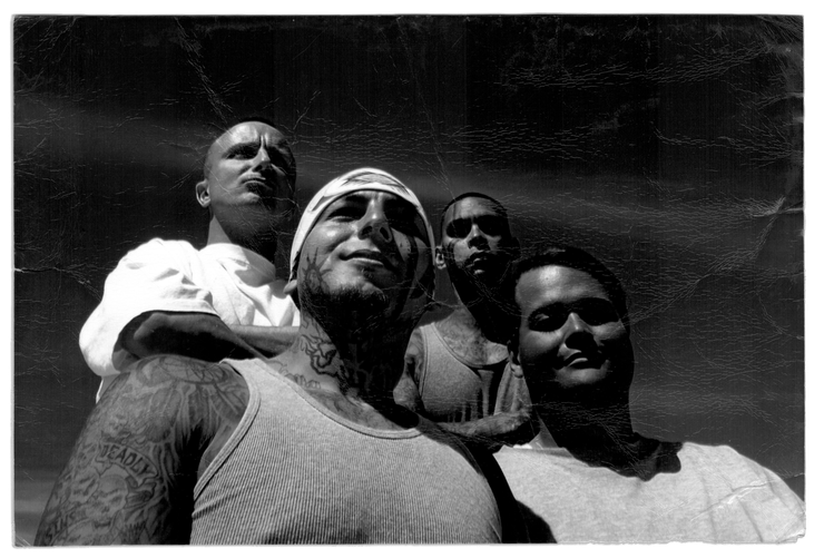 In a black and white photograph, Grace Pinson, a trans Latina with short dark hair, stands outside in the sun with three men. The person to her left wears a tank top and has tattoos on his face, neck and arms. Behind her stand two men. The one on the right wears a white T-shirt and is bald. The one on the left has tattoos on his chest and arms and has close-cropped, dark hair. 