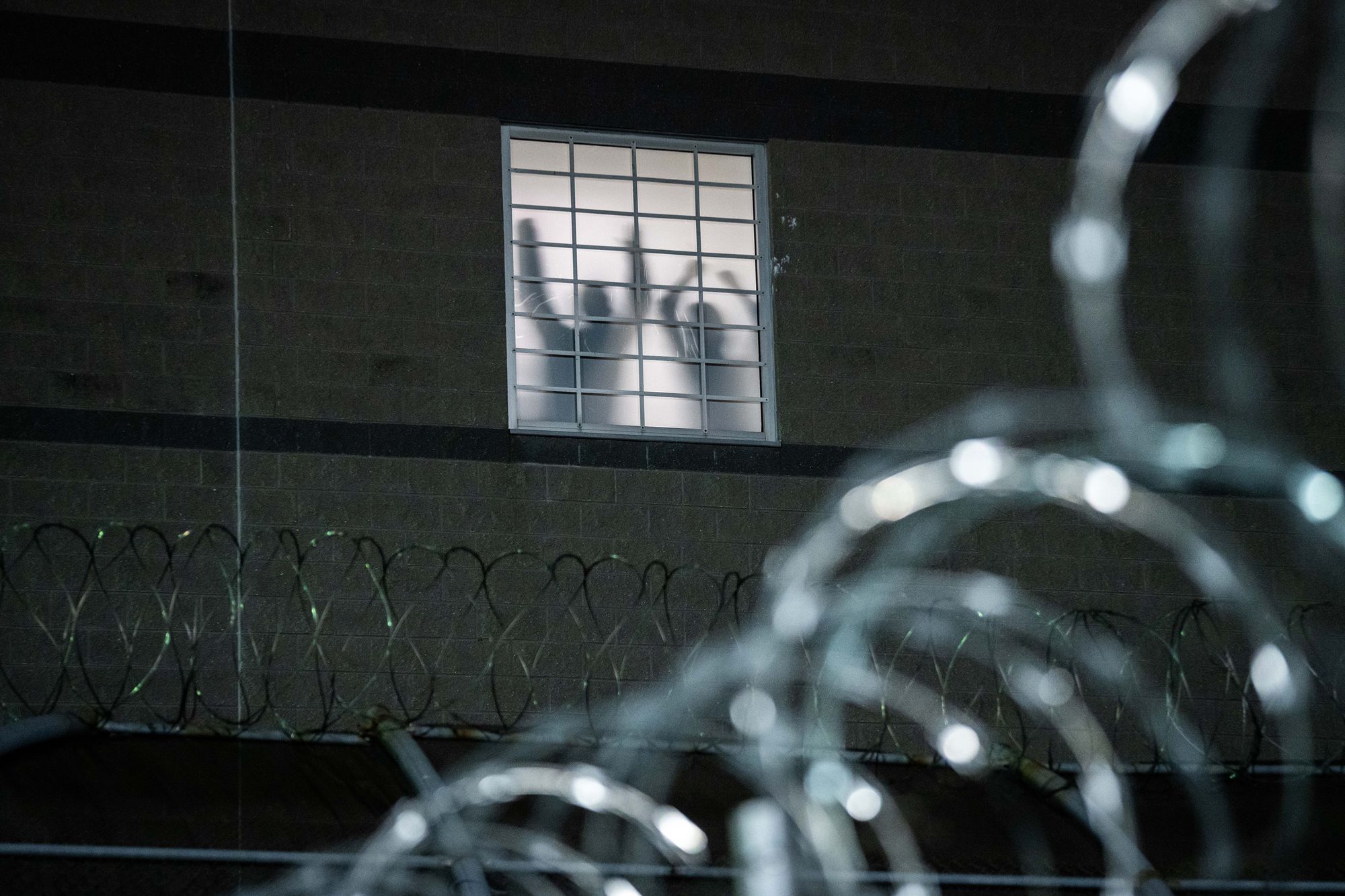 A photo shows two people’s silhouettes visible through a window of a federal immigrant camp. Their arms are raised. Below the window is barbed wire, and in the foreground is more barbed wire, blurred and close up.