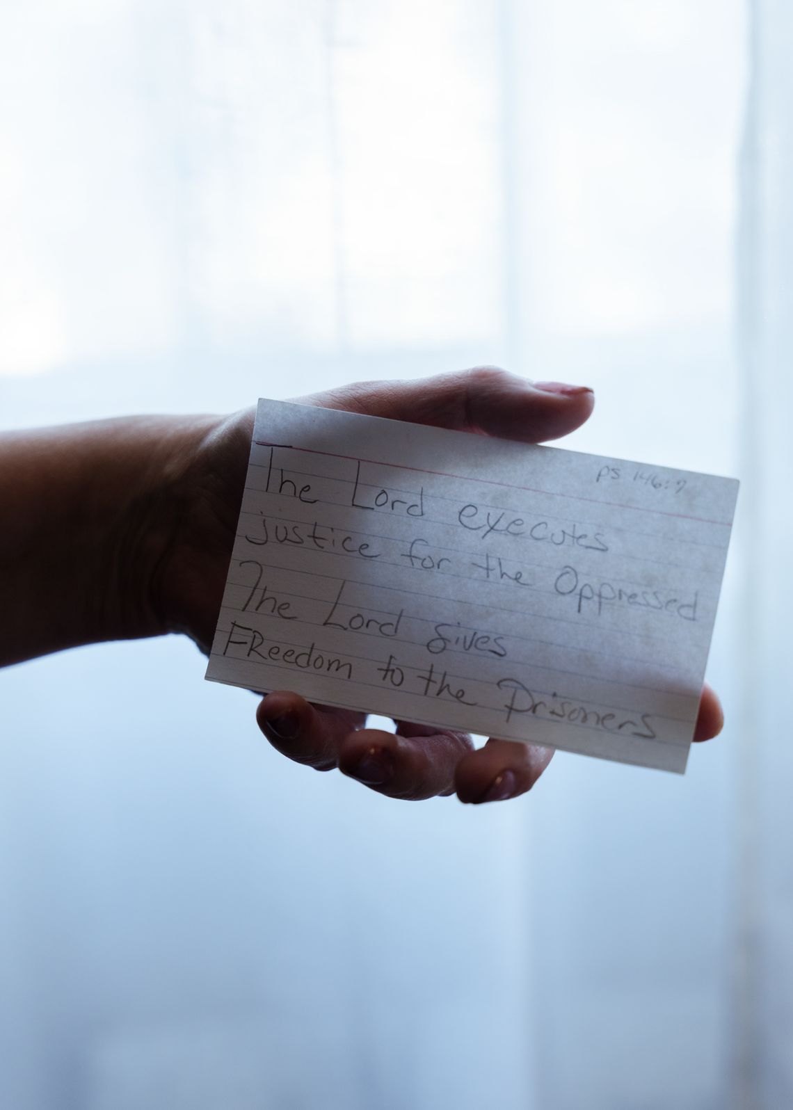 A photo shows a White woman’s hand holding up an index card which has “The Lord executes justice for the oppressed. The Lord gives freedom to the prisoners.” The background is a white window curtain with sunlight shining through it. 