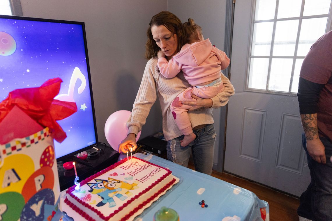 While holding a toddler, a White woman with brown hair lights a birthday candle on a cake with cartoon dog characters.