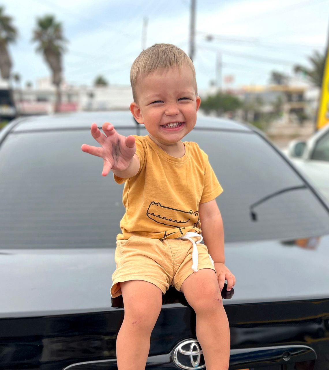 A photo shows a boy with blonde hair and light-toned skin, smiling as he sits on the back of a black car.