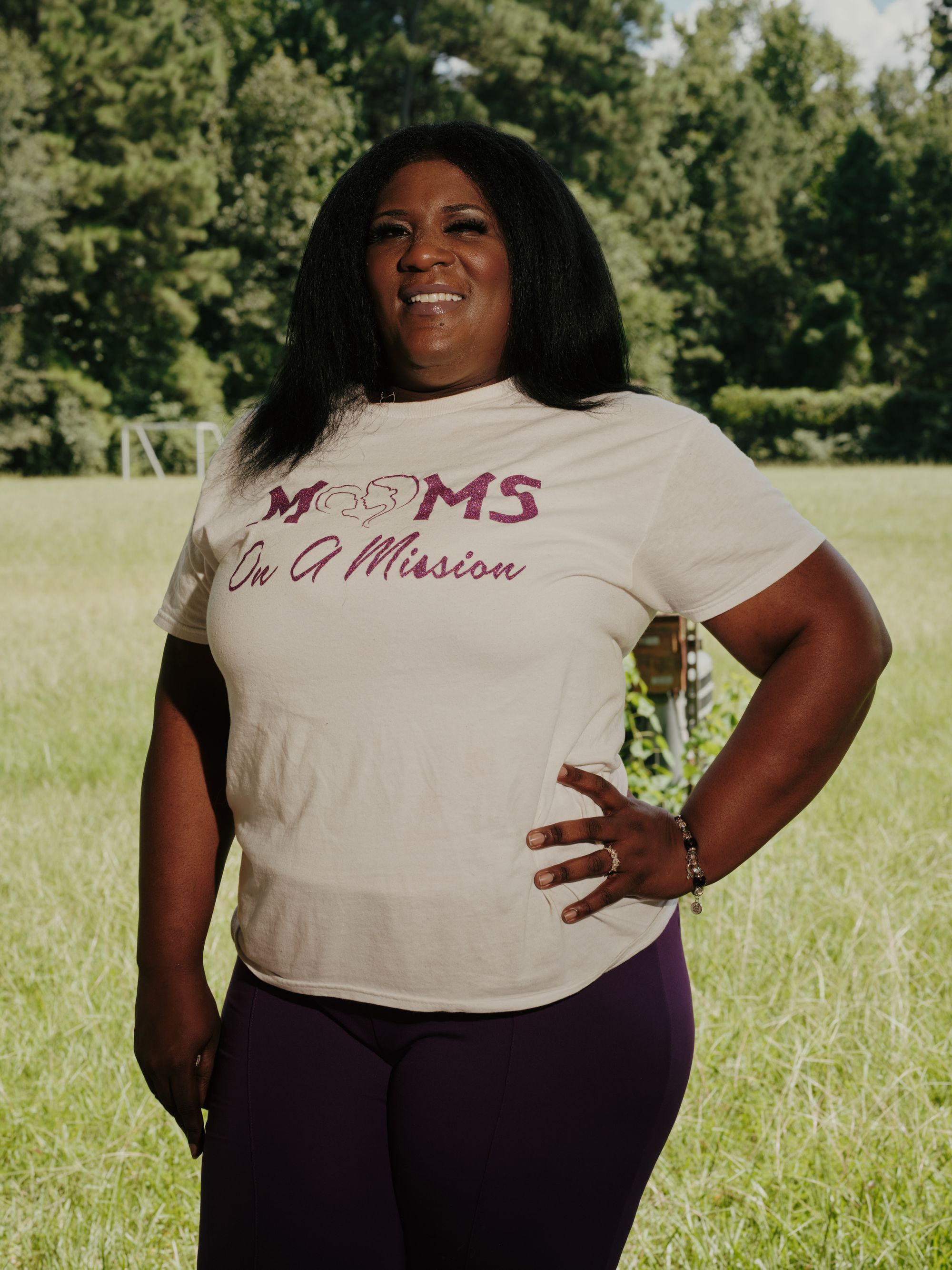 Martha Tyler, a Black woman wearing a white T-shirt that reads “Moms on a Mission,” poses outdoors for a photo while smiling. Trees and grass can be seen behind her.