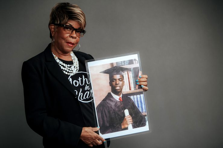 Dr. Dorothy Johnson-Speight, a woman with dark brown skin and wearing a black blazer and shirt, stands in front of a gray studio background. She holds an image of her son Khaaliq Jabbar Johnson, who is wearing a graduation cap and gown. 