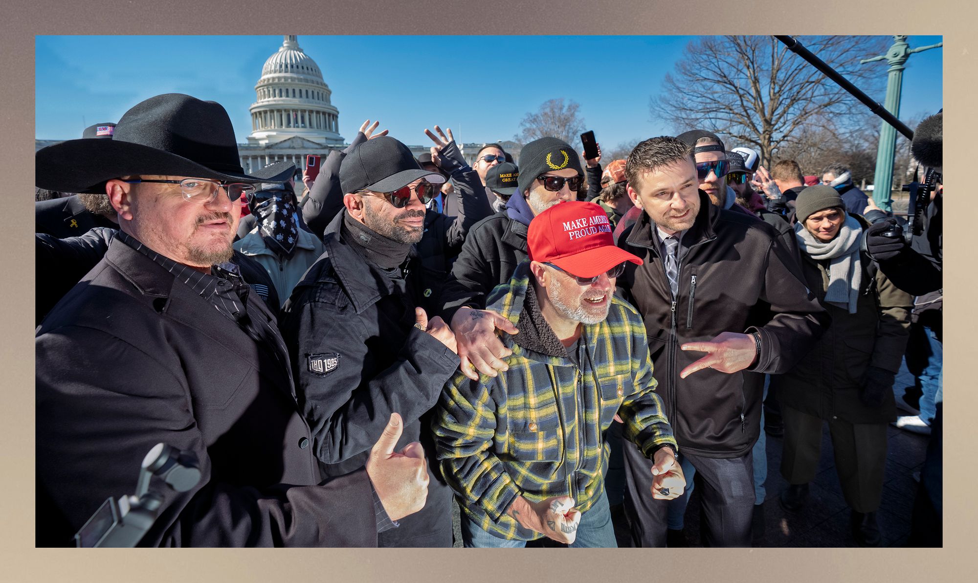 A group of men, including from left, Stewart Rhodes, a man with light-toned skin, wearing a black hat and glasses with a patch over his left eye, and Enrique Tarrio, a man with medium-toned skin, wearing sunglasses and a black baseball cap, stand outside in Washington, D.C. Both have their thumbs up. 