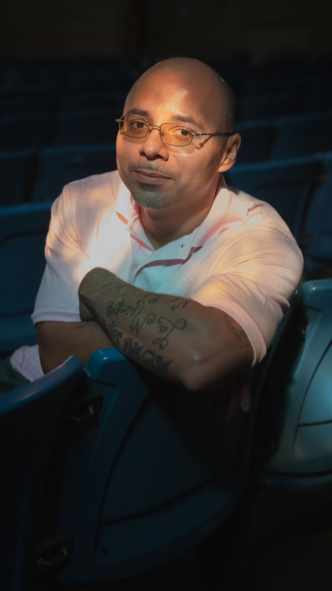 A bald Latino man wearing glasses sits in a theater chair in the shadows, his tattooed forearm perched on the seat. The light shines on his face, which bears a small smile. 