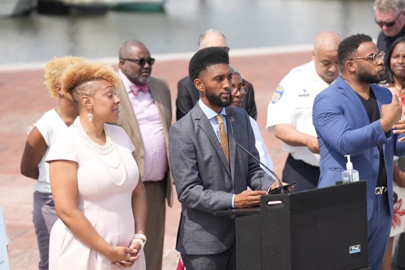 The mayor of Baltimore, a Black man in a gray suit and a gold-colored tie, stands at a podium during a news conference in May while a Black woman in light-colored dress stands beside him with her arms clasped in front of her. A mixed group of people is behind them.  