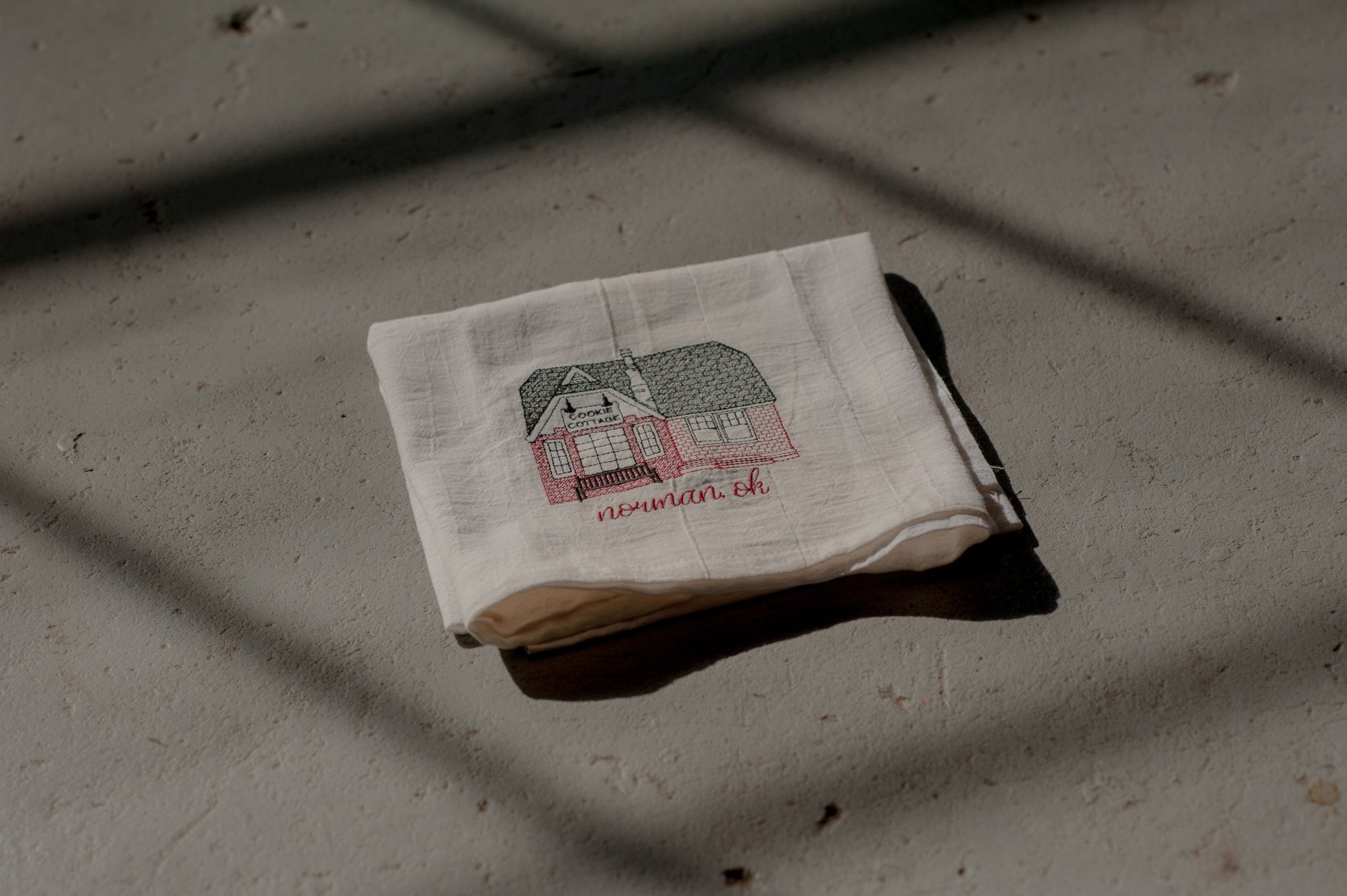 A white hand towel with an illustrated depiction of the Cookie Cottage building. The towel is lying on a dark gray floor, framed by the shadow of the window panes. 
