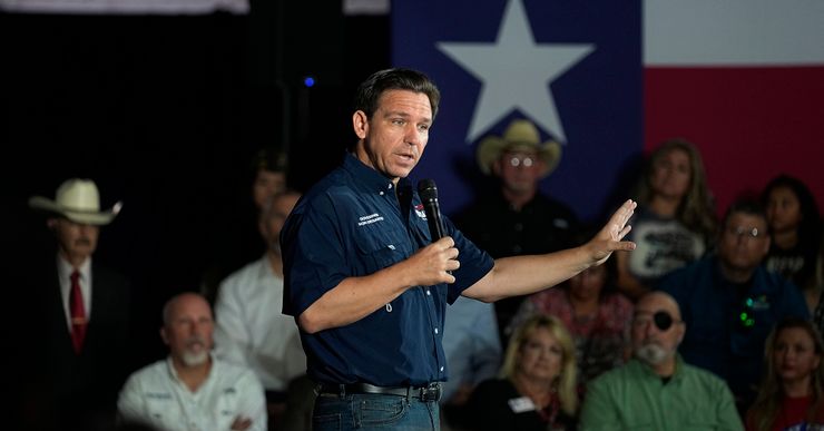 A White man, wearing a short-sleeved blue button up shirt and jeans, holds a microphone as he speaks to a crowd. Several people are seated in the background. A Texas flag hangs from the wall. 