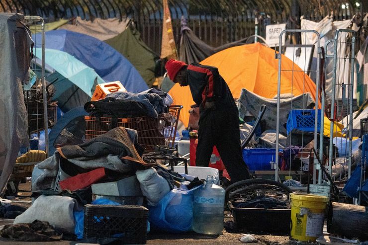 A man wearing a red beanie and a black and red jumpsuit looks down as he stands in the middle of a homeless encampment. 