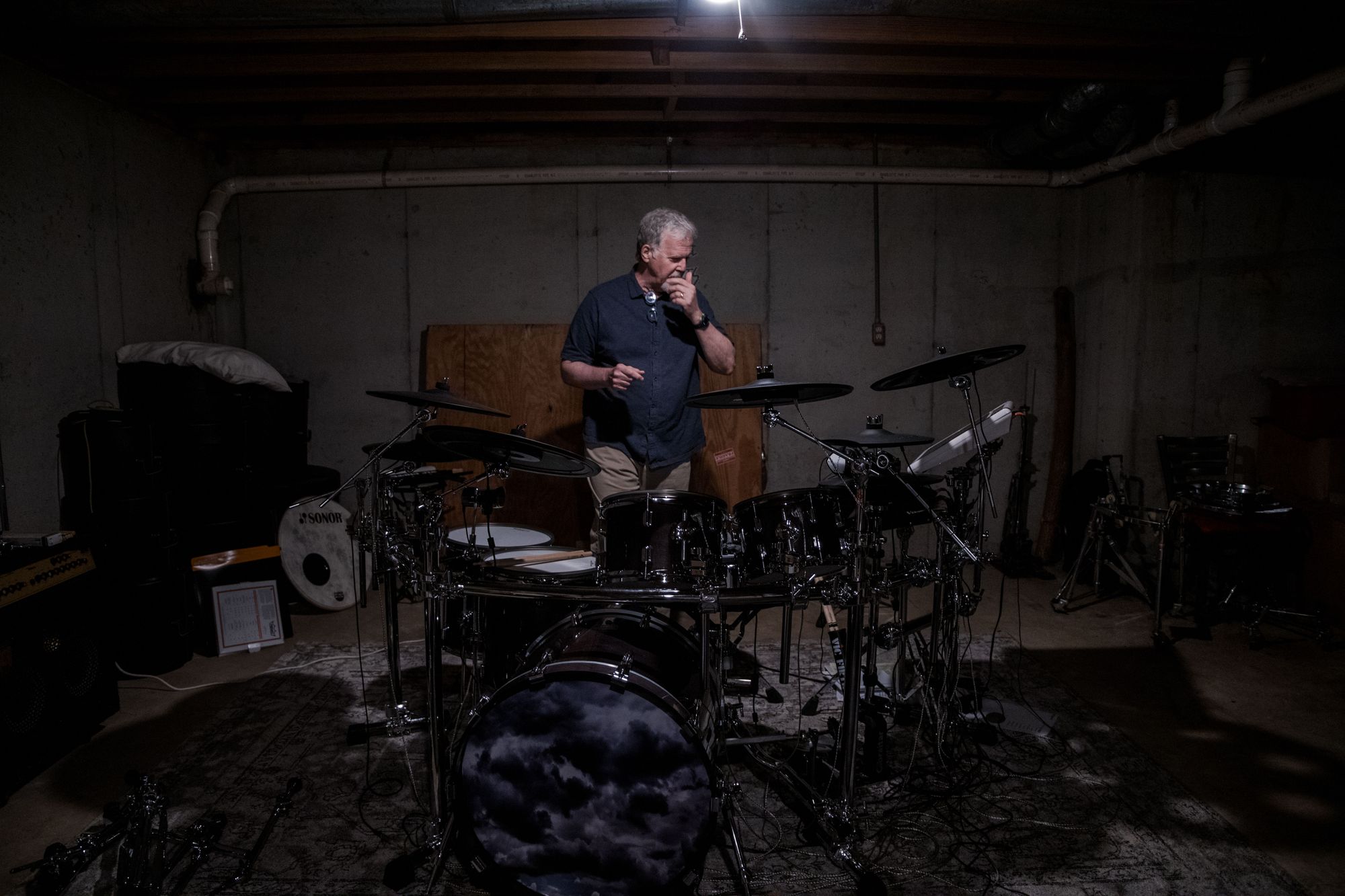 Belzley, a White man with gray hair and a goatee, wears a blue button-down shirt and khakis, and stands behind a large drum kit.