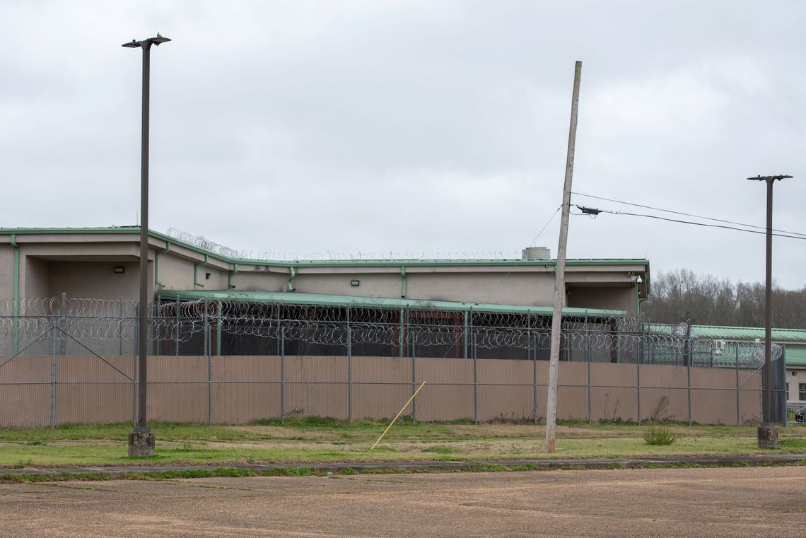 A photo of a beige building with a light green roof behind a barbed wire fence.