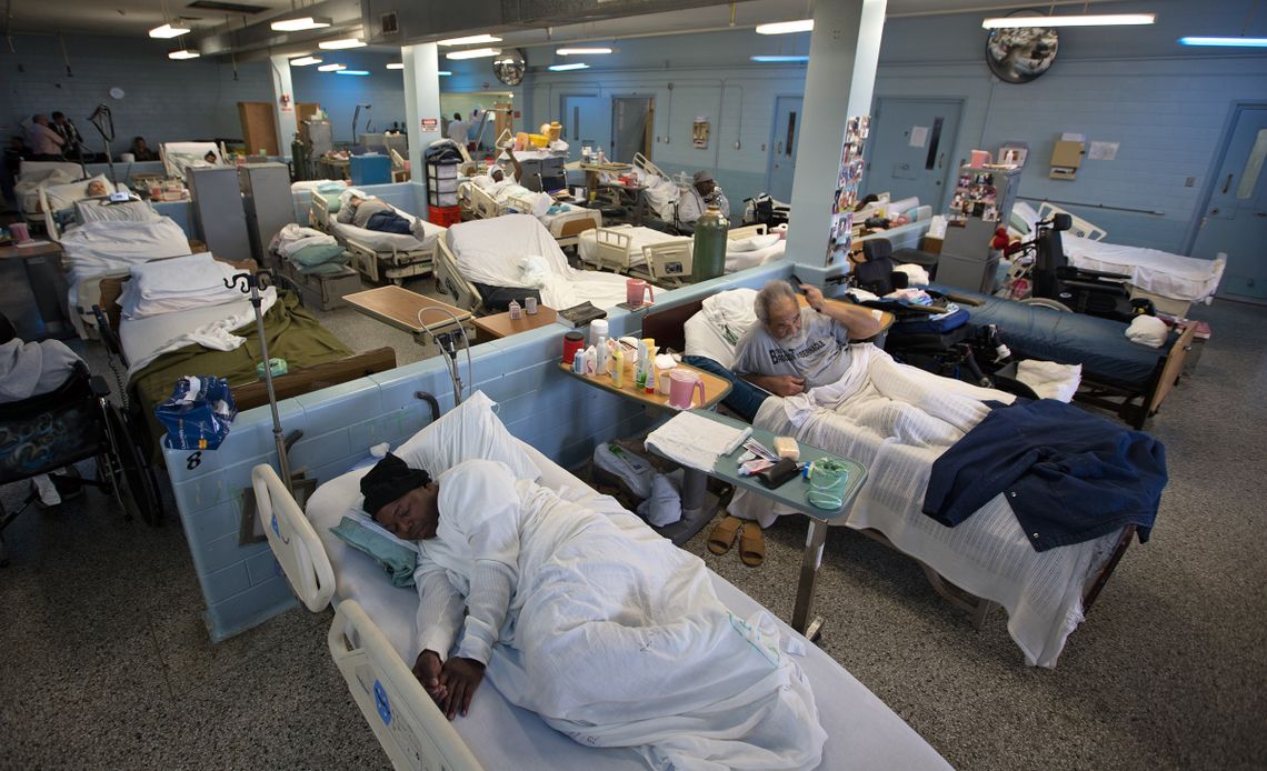 Elderly people are lying in beds in a geriatric ward. A man with white hair and a white beard is combing his hair. 