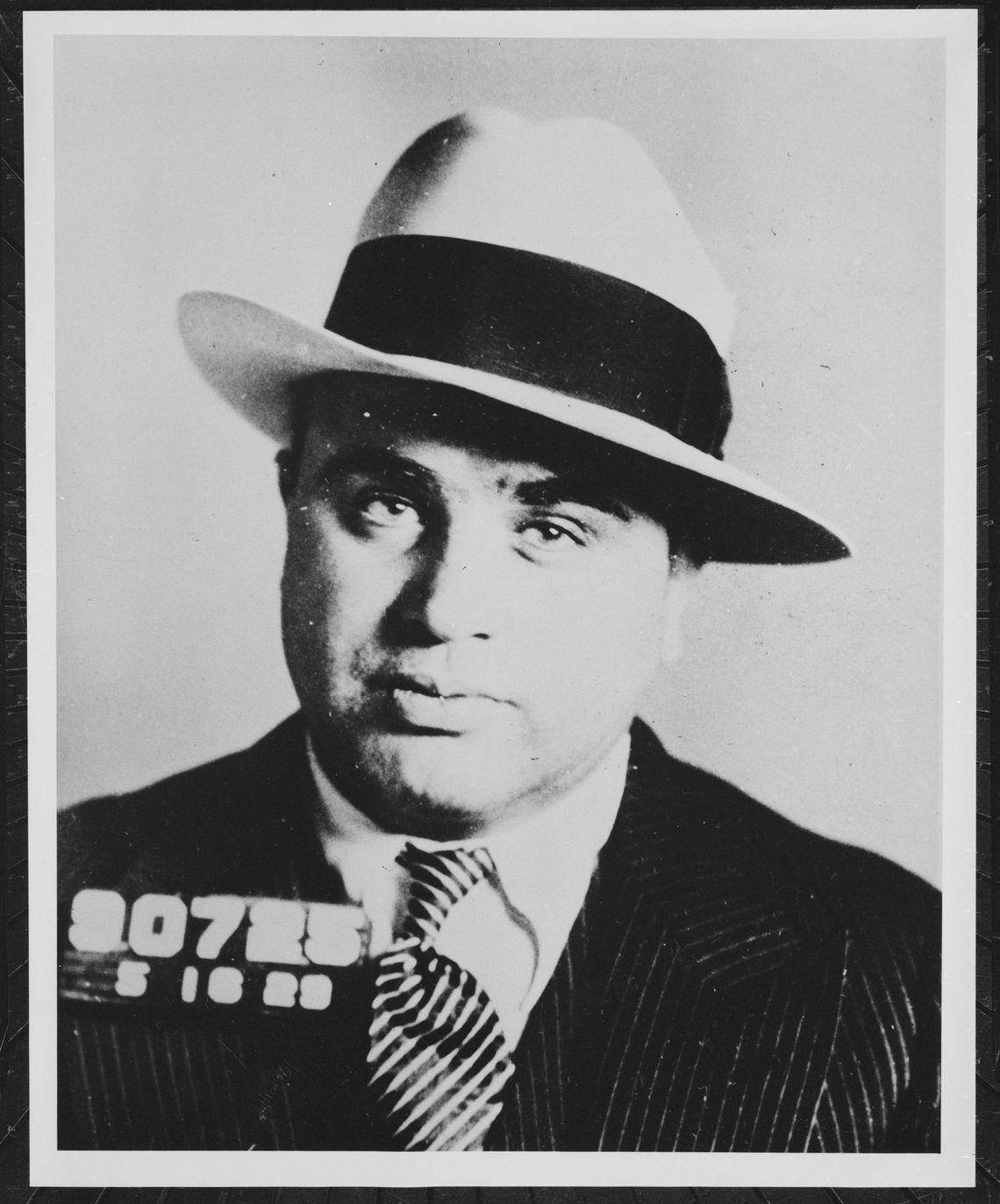  A black-and-white photo shows a White man wearing a suit and tie, and a light-colored hat with a black band.