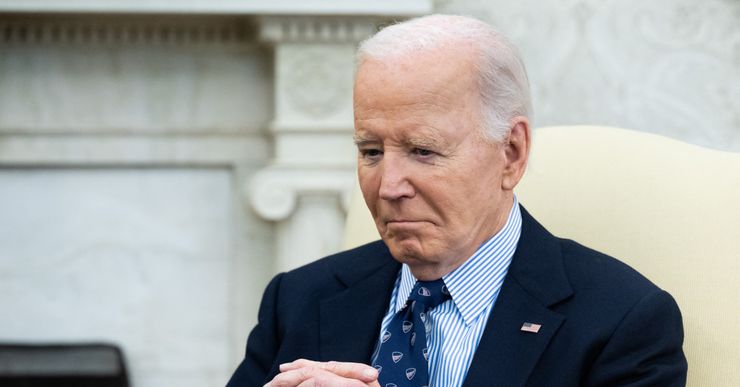 U.S. President Joe Biden sits in a white chair in a navy suit with his hands clasped in front of him.