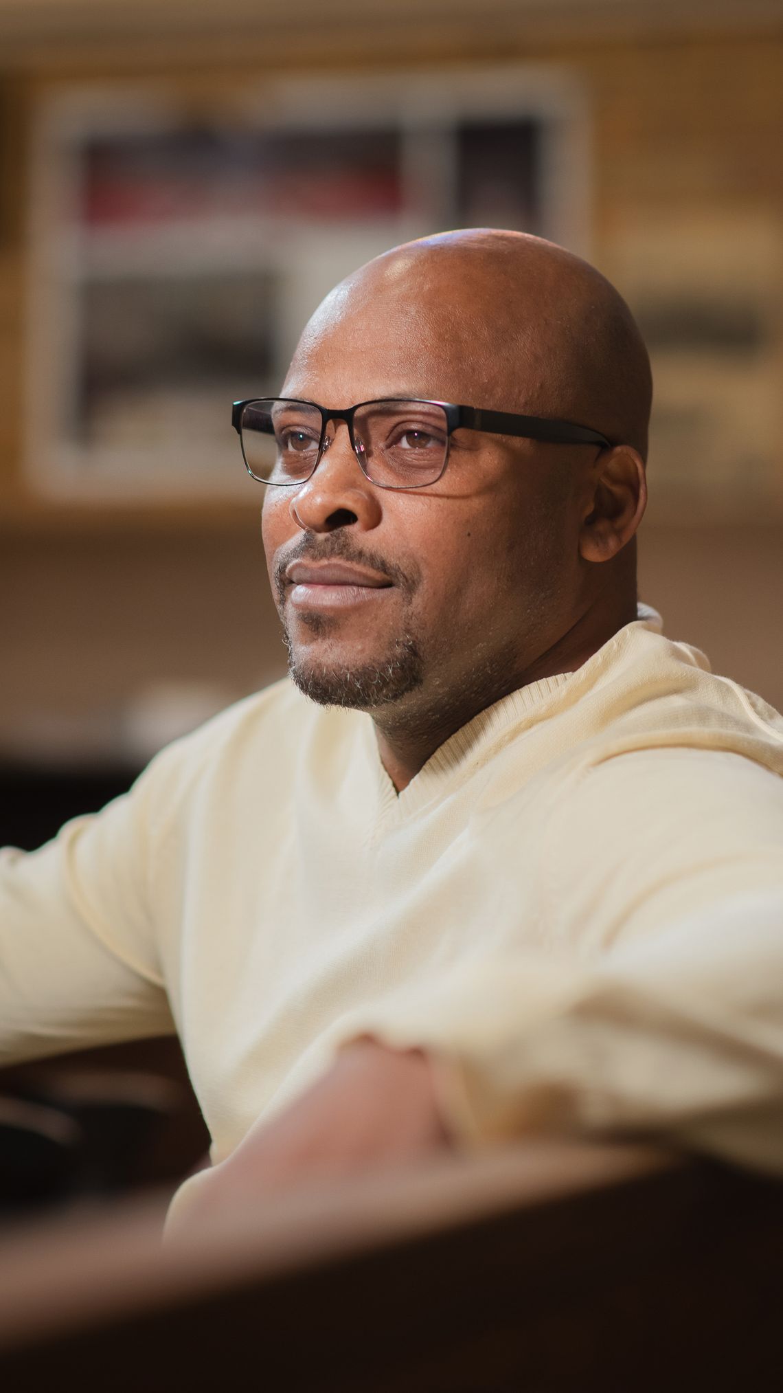 A bald Black man with a goatee, wearing glasses, sits in a church pew wearing a cream-colored sweater. 