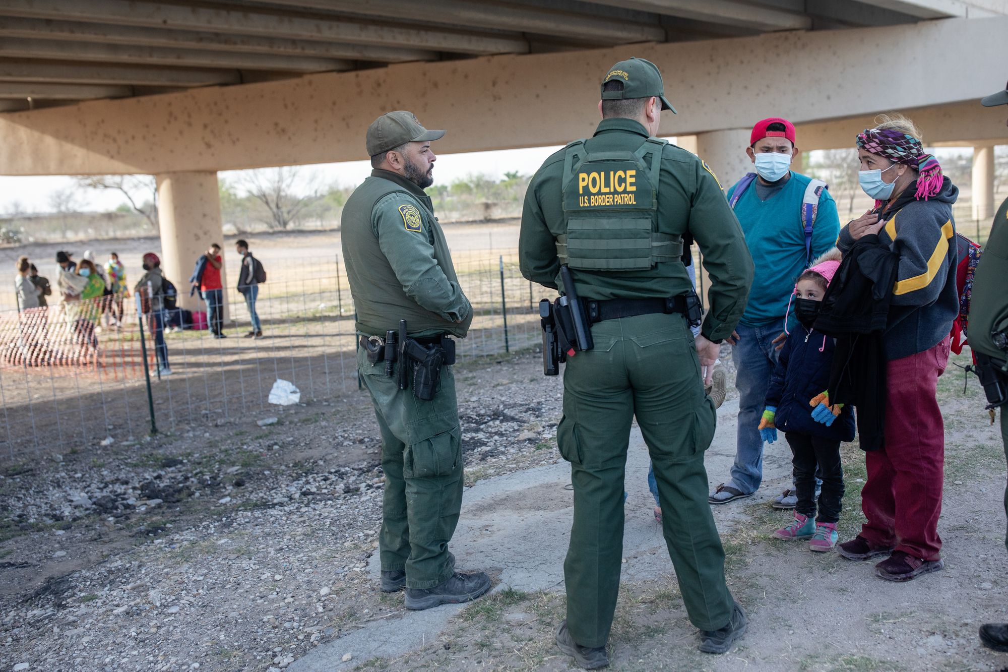 On the right, two adults and a child wearing face masks face two Border Patrol agents wearing green uniforms. They, and a group of people in the background, are standing under a bridge. 