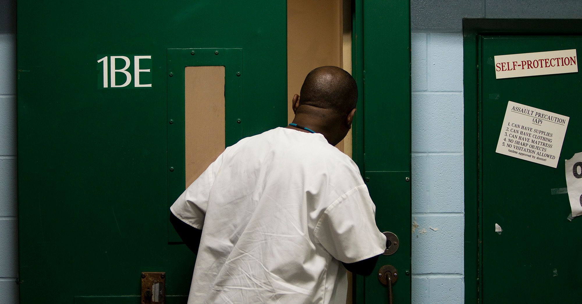 A Black medical ward assistant in a white uniform looks through an open door. 