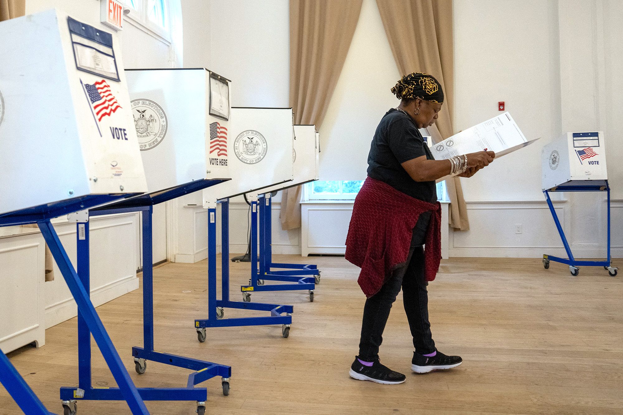 A Black woman, wearing a black shirt, red sweater and black pants, holds her ballot at a polling place. 