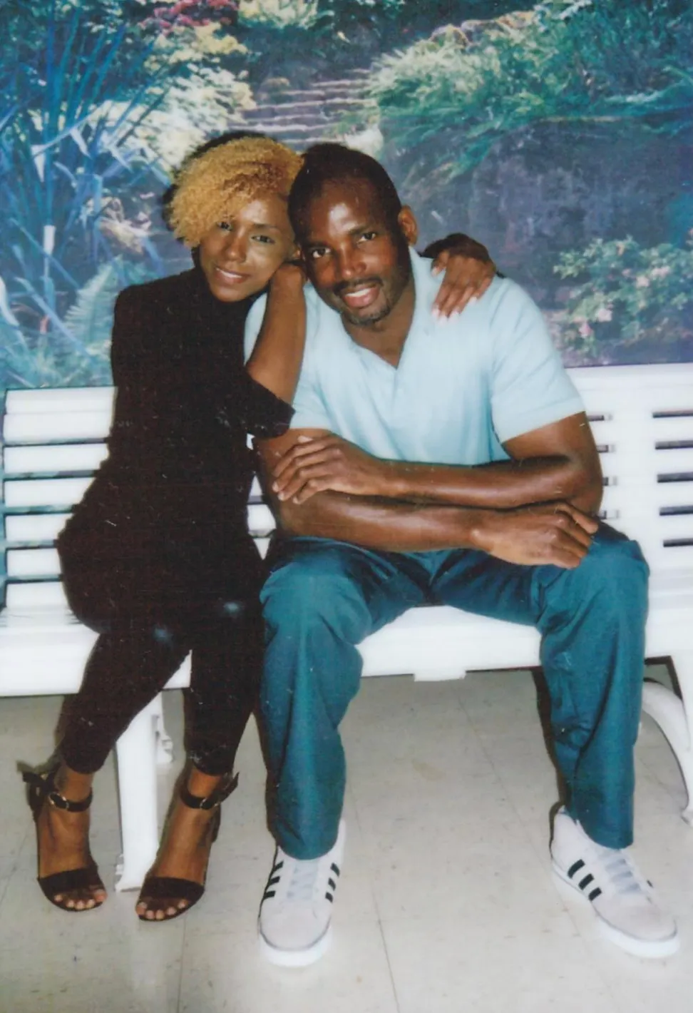 A photo shows a Black man wearing a pale blue shirt and teal pants sitting with a younger Black woman with short blonde hair, wearing a black shirt and pants. 