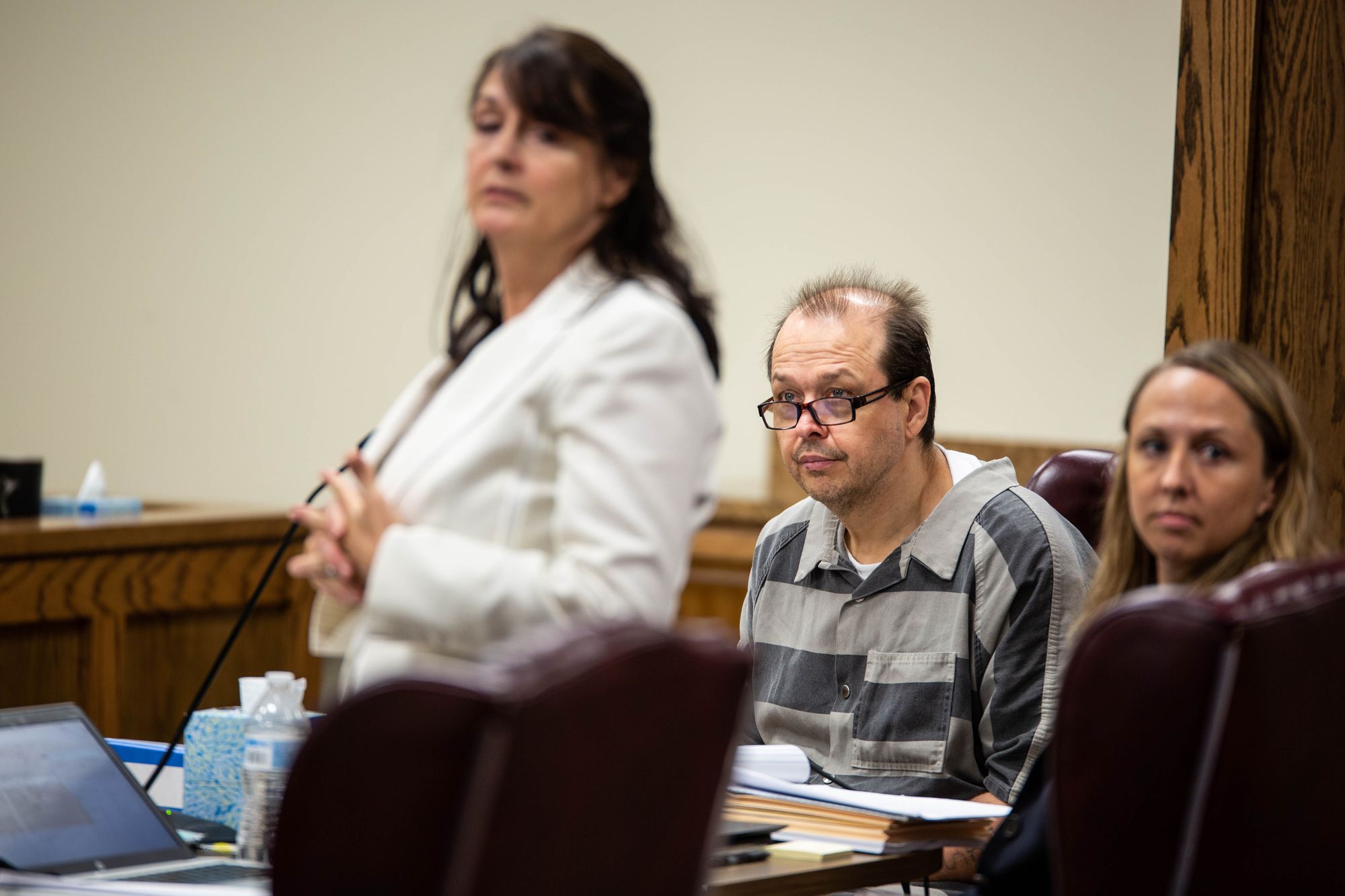 In a courtroom, a White man sits at a table peering over thick-rimmed black glasses. He is dressed in a gray-striped shirt. To his left is a woman standing in a white suit jacket and to his right is another woman, seated at the table next to him.