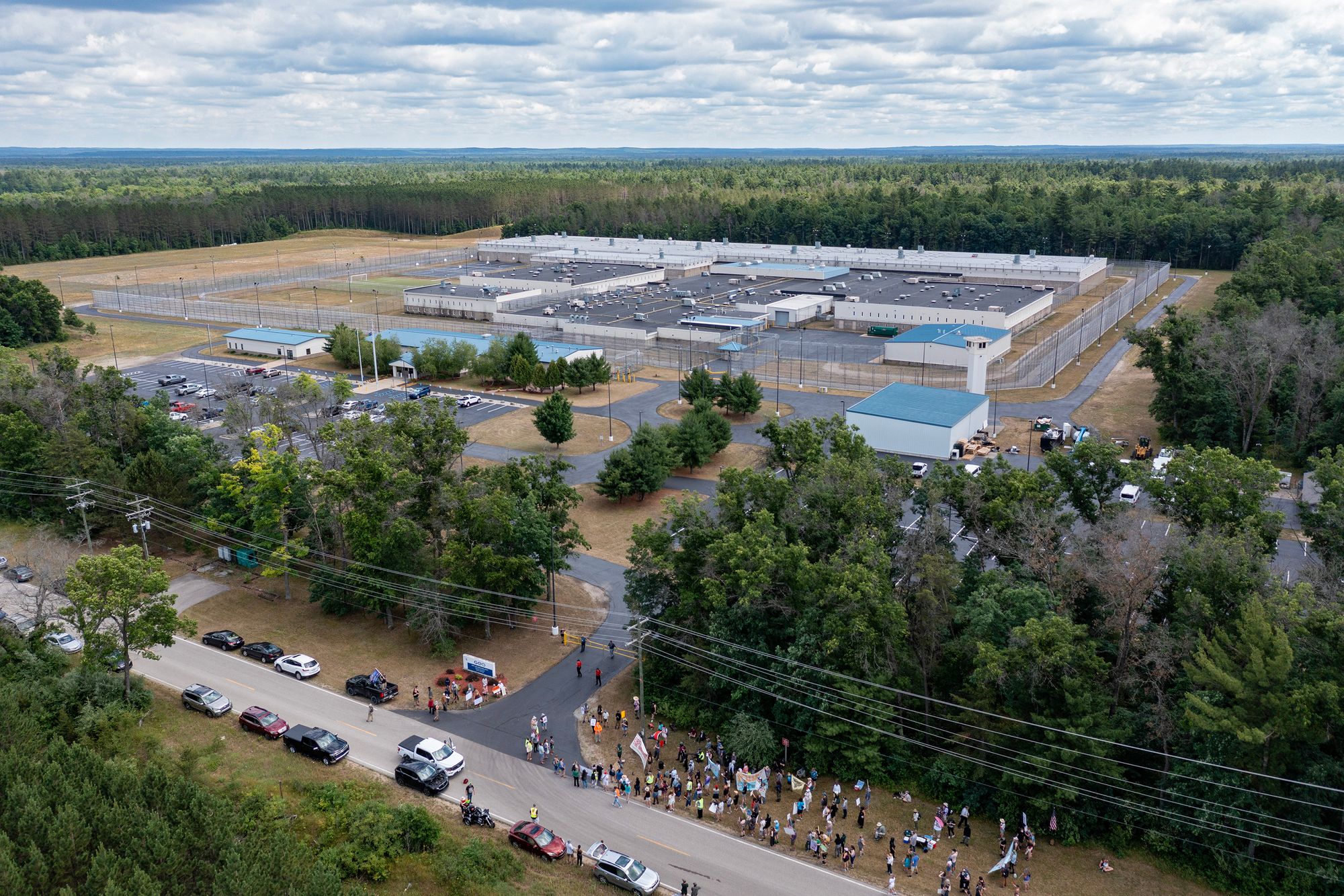 An aerial photo shows a prison facility composed of over half a dozen buildings, with trees surrounding much of the property. In the foreground, there are activists gathered with signs and banners near the facility.  