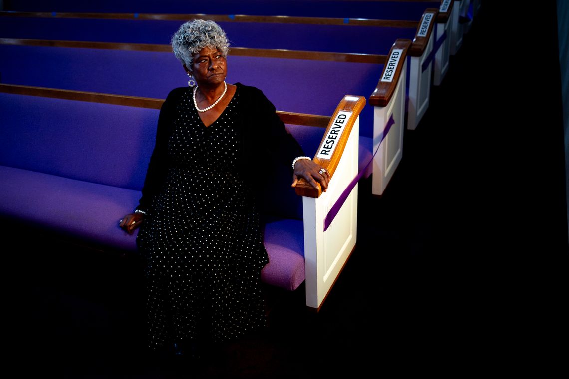 A Black woman with a long dark dress and short curly hair looks out a window while sitting in a purple church pew.