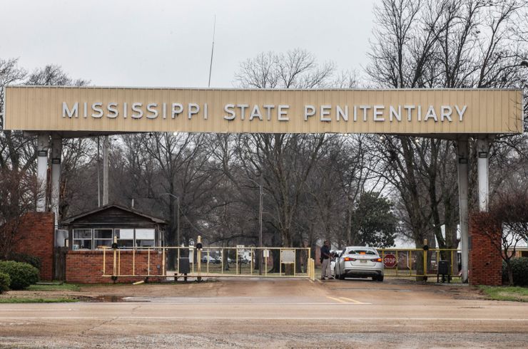 A photo shows a silver car going through a prison checkpoint entry with a large sign that reads “Mississippi State Penitentiary.” Bare trees are visible in the background beyond the entrance.  
