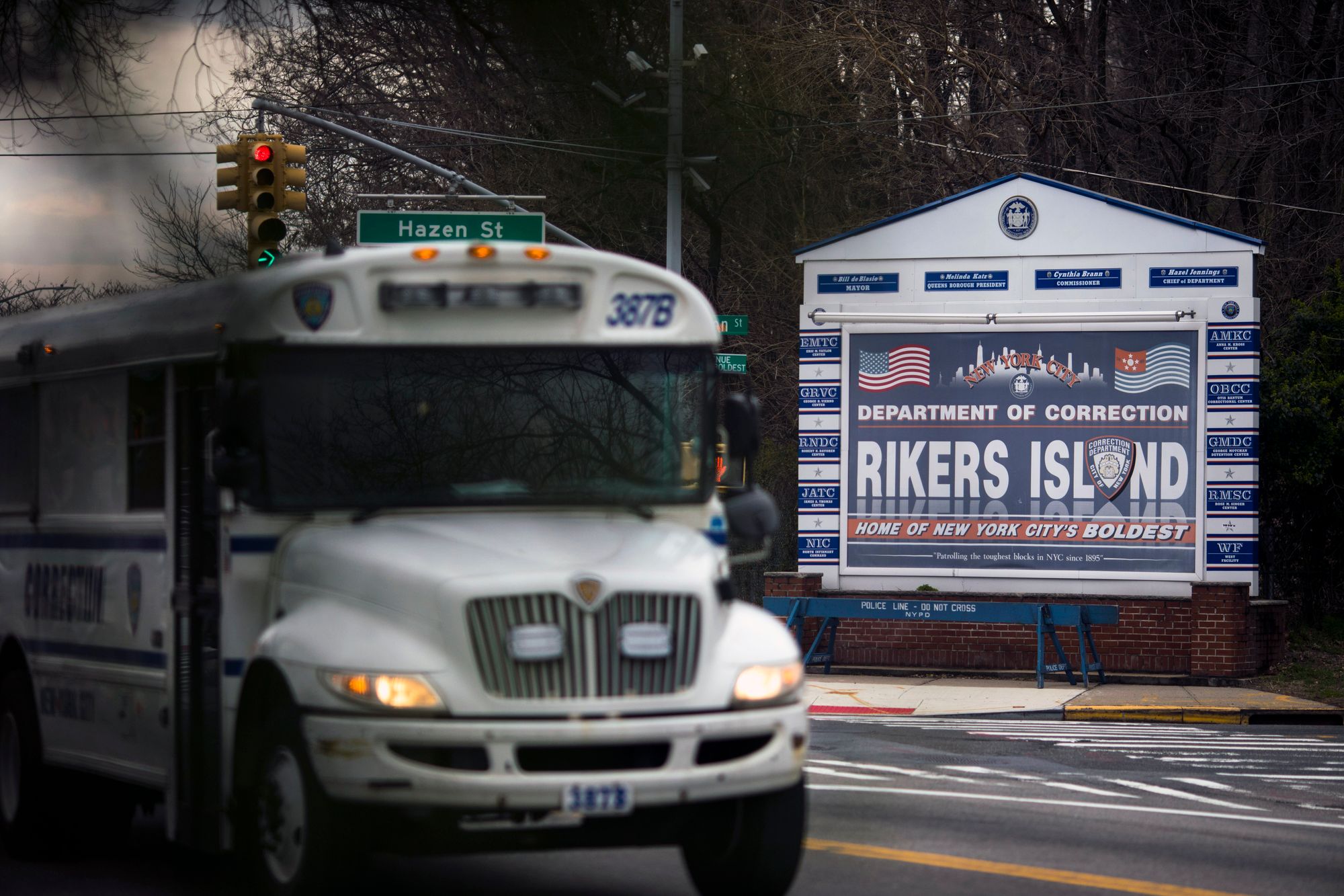 A white bus drives in front of a sign that says "Department of Correction: Rikers Island."  