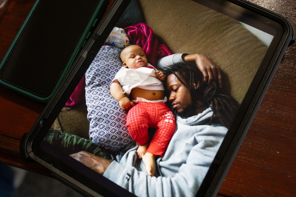 A photo shows a tablet, and on the screen of the tablet is a photo of a Black man wearing a gray sweatshirt lying next to a Black baby wearing a white T-shirt and red patterned pants. The baby is resting on a blue patterned pillow. 