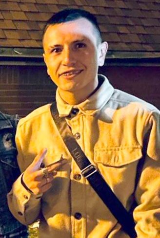 A light-skinned Latino man, wearing a white buttoned shirt and black arm sling, smiles for a portrait while holding up a peace sign. 