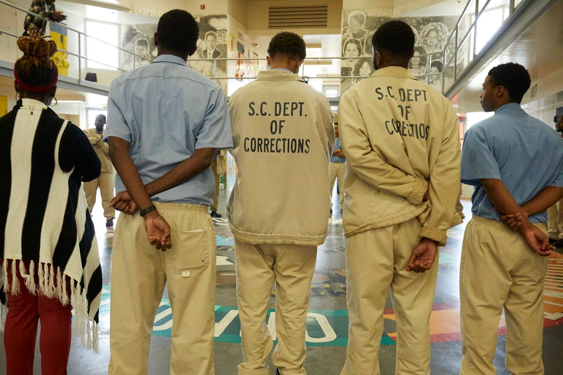 Five people stand in the common room of the Turbeville Correctional Institution with their backs to the camera. A woman stands on the left, wearing a vertically striped black and white sweater. Four Black men with short cropped hair, two of whom are wearing jackets that read “S.C. Dept. of Corrections” stand to the right of her.