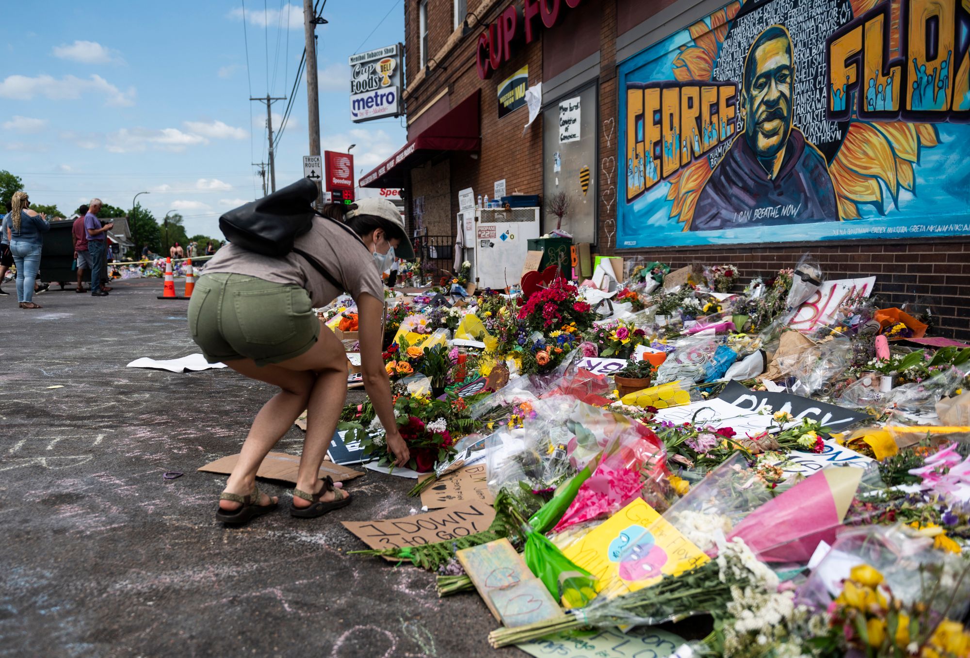 A woman with light-toned skin crouches down to place flowers at a memorial. Hundreds of flower bouquets, cards and signs are visible on the ground, and an orange and blue mural showing George Floyd with a backdrop of a sunflower is on the wall.  