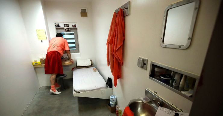 A man wearing an orange and red prison uniform looks out the window of his solitary confinement cell. 