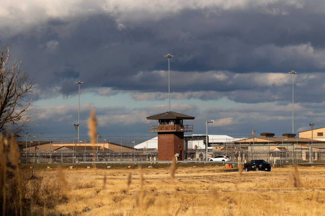 A photo shows a prison that has a red-brick lookout tower. Chain link fencing with barbed wire on top is behind the tower. In front of the tower is a field of yellow grass. The sky is mostly filled with dark clouds, with patches of pale blue. 