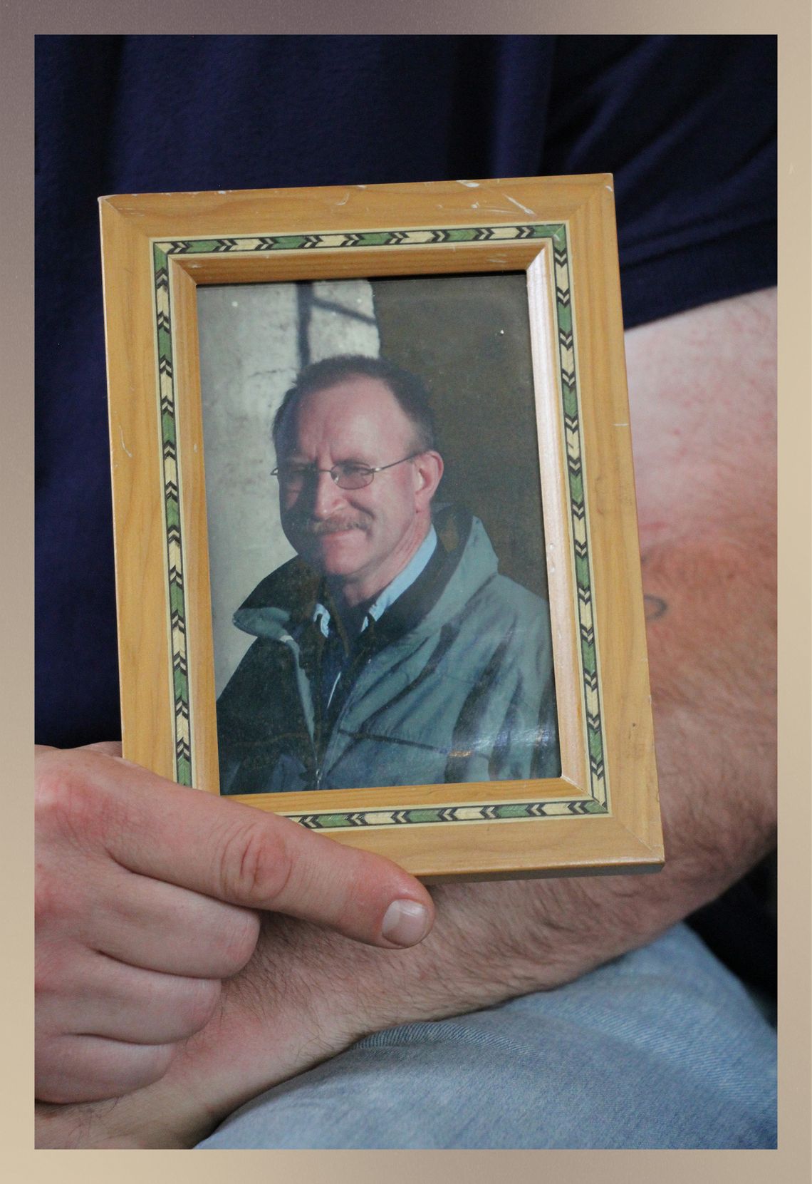 A person’s hand holds a framed photo of a White man with a mustache, who is wearing glasses, a green jacket and a light blue button-down shirt. 