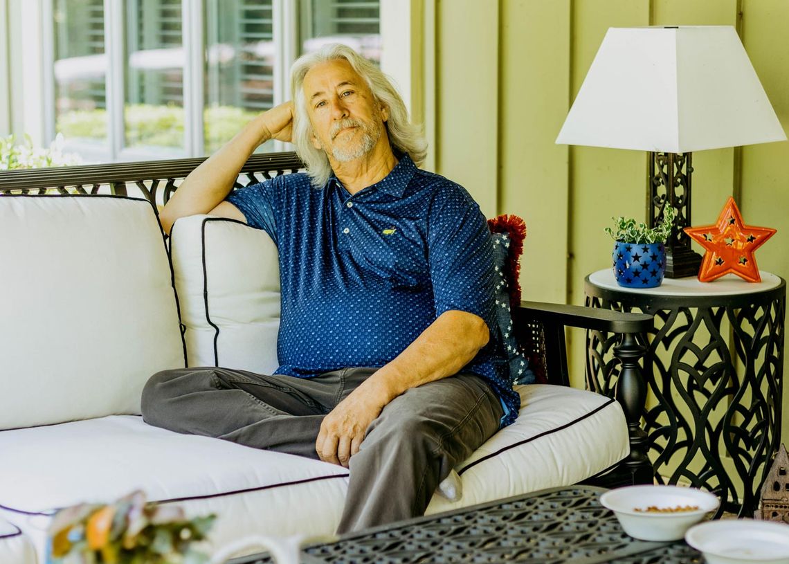 A White man with white hair wearing a blue shirt sits on a couch.