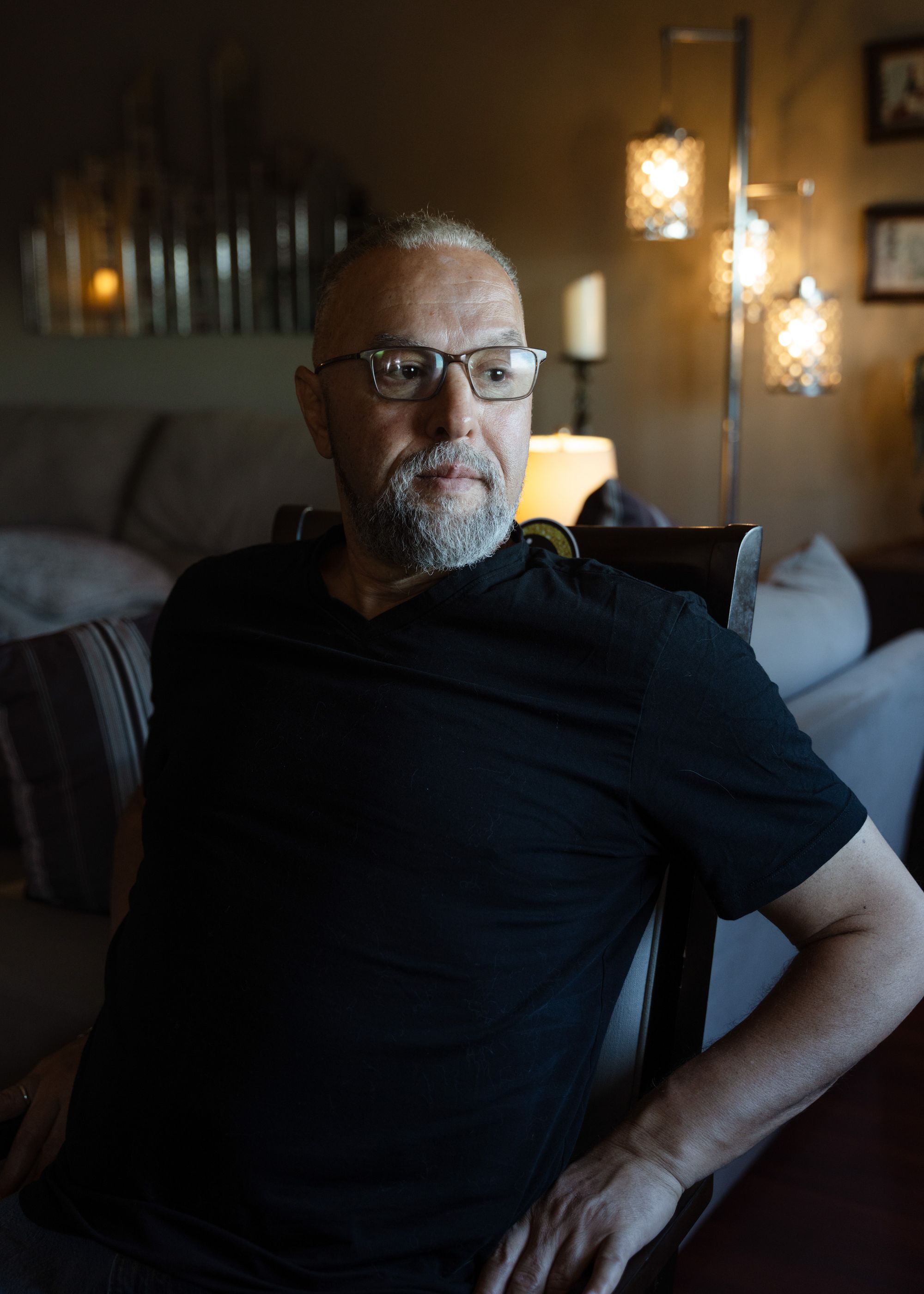 A photo shows Latif Hafraoui, a man with medium-toned skin. Latif has a gray beard and glasses and is wearing a black t-shirt. He is sitting on a chair at his home. A couple of sofas are behind him. 