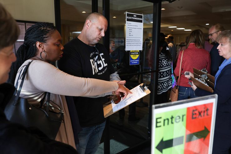 A White man wearing a black T-shirt, a gray long-sleeved shirt and blue jeans, holds a clipboard with a voter registration form. A Black woman wearing a light gray cardigan and holding a black purse, points at the registration form as they stand at the entrance of a building. People are standing in front of them in a line. 