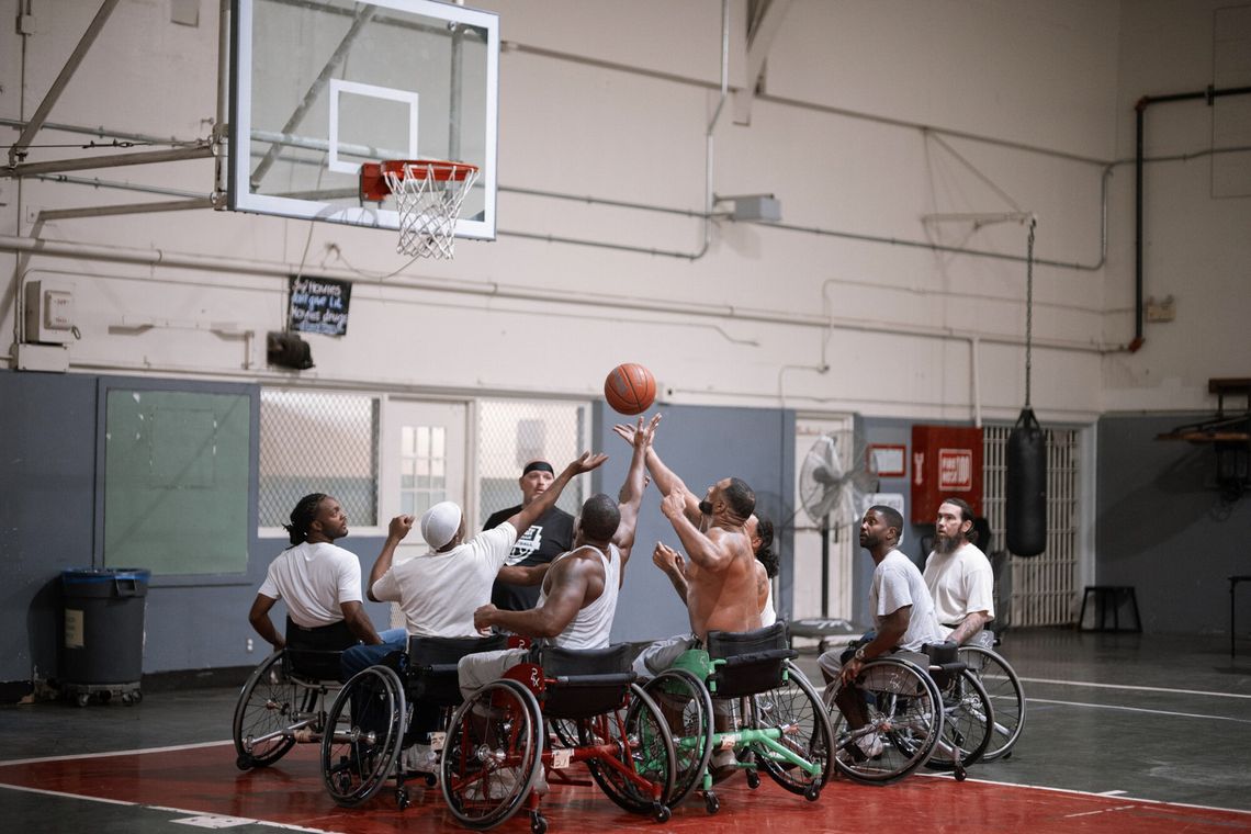 A photo shows a group of wheelchair basketball players all reaching up simultaneously for a basketball while in an indoor court. 