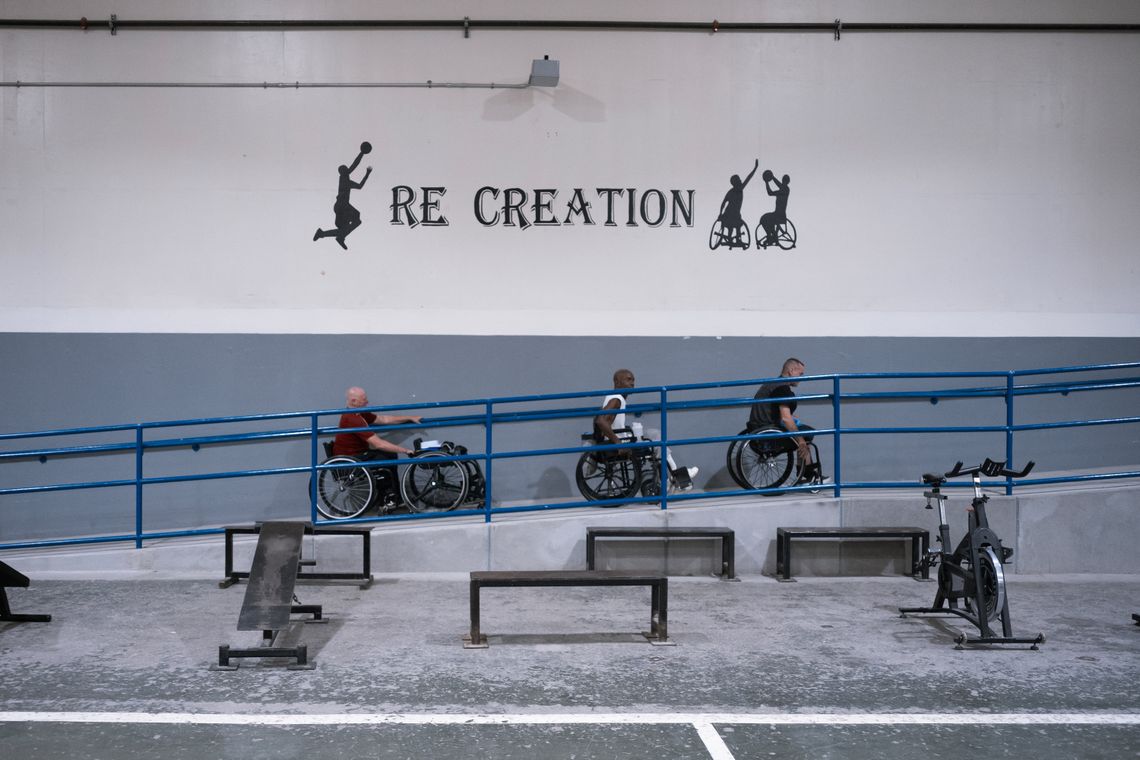A photo shows three people in wheelchairs wheeling themselves up a ramp. On the wall to their right is painted the word Re creation” with some small painted silhouettes of people playing wheelchair basketball. 