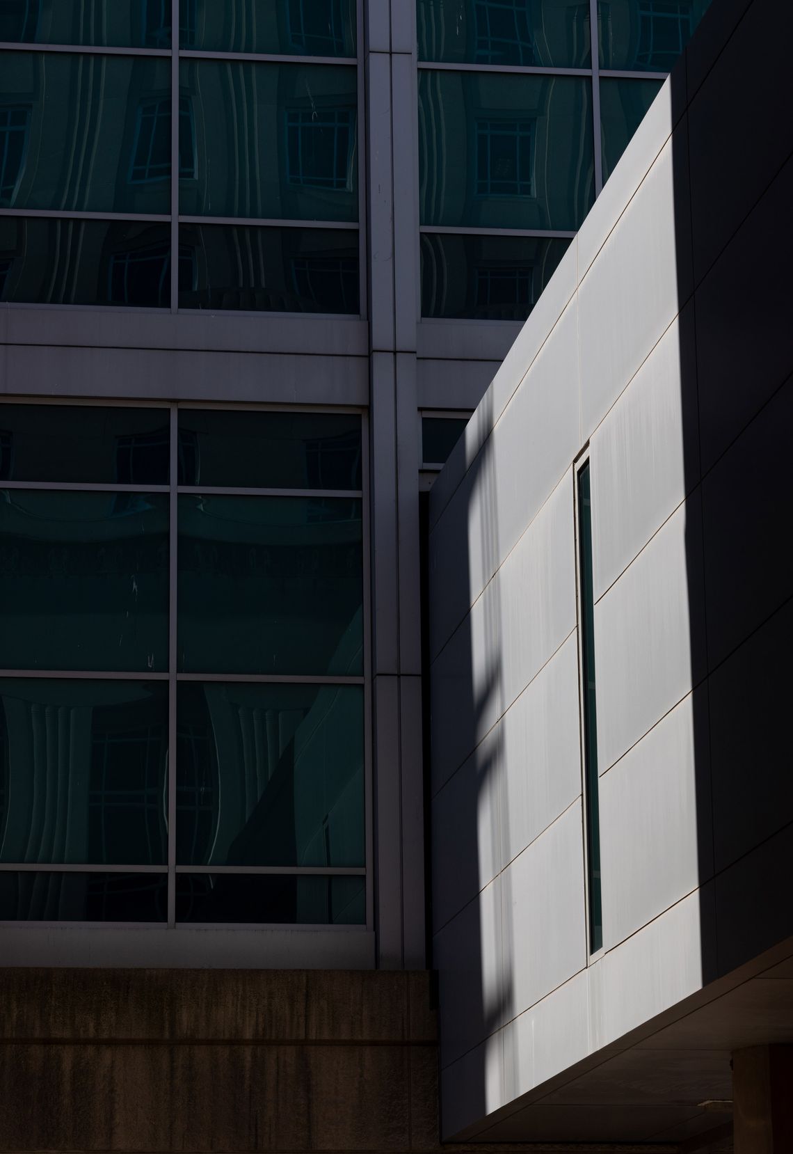 A photo shows a close view of the outside of a jail building. One side, in shade, has blue window panes. Another side, in harsh sunlight, shows a wall of concrete with a thin, vertical window.